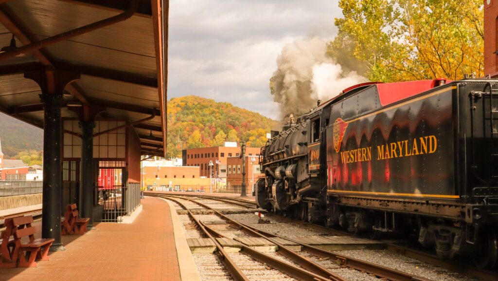 A Western Maryland Scenic Railroad train at a station with a mountain with fall foliage in the distance