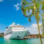 Cruise ship anchored in light turquoise water off the coast of a small Caribbean island with palm trees in the forefront