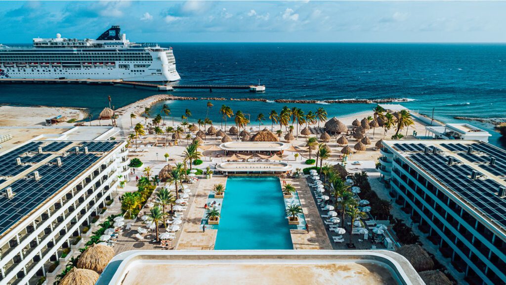 Resort flanked by two buildings with a pool in the middle, facing the beach and Caribbean Sea with a cruise ship in the background