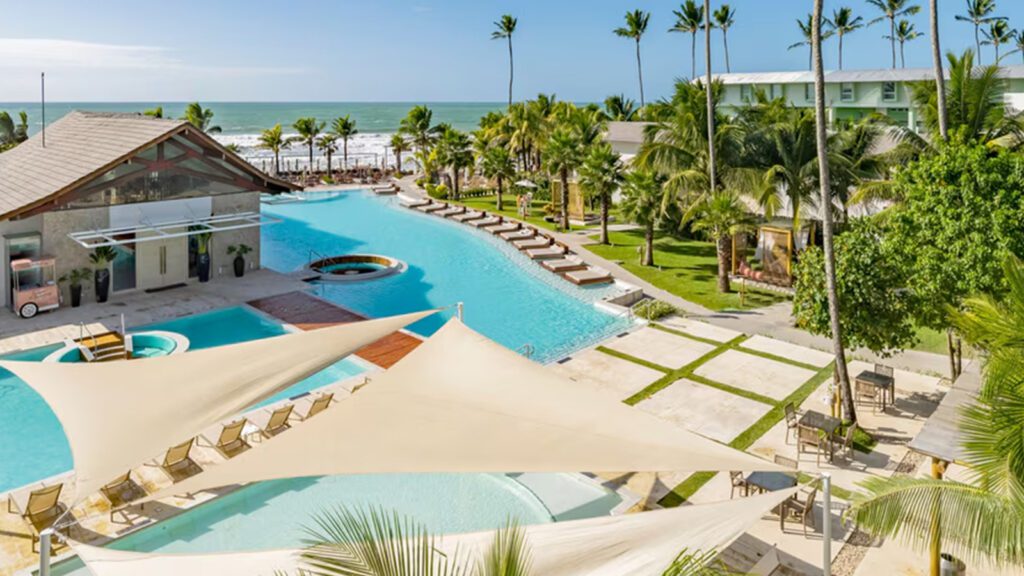 Swimming pools surrounding a small building amidst a grassy area with palm trees and the beach in the background