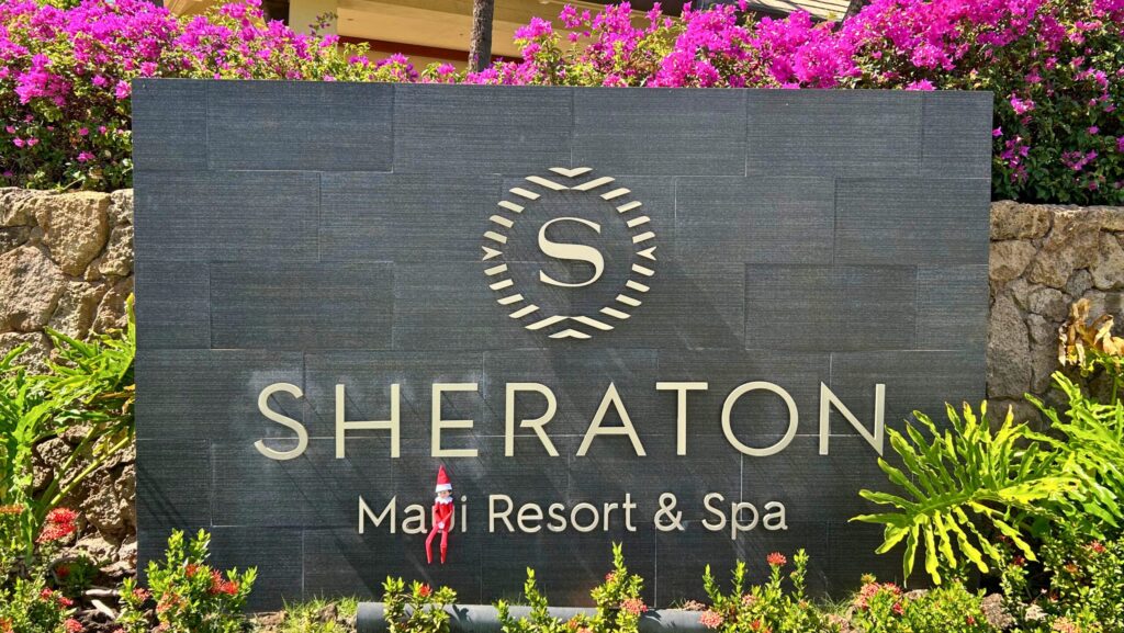 An Elf on the Shelf sitting on a sign surrounded by tropical greenery at Sheraton Maui Resort & Spa