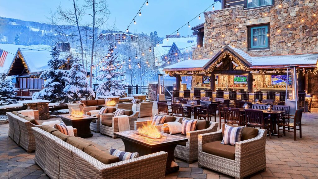 An outdoor bar and lounge area with a firepit and string lights and a snowy mountain in the background at The Ritz-Carlton, Bachelor Gulch