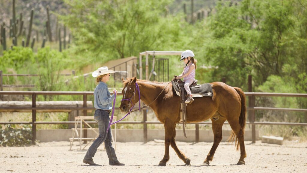 A girl in a helmet riding a horse led by a woman in a cowboy hat at Tanque Verde Ranch