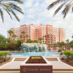 Pool with waterfall feature framed by two palm trees, and pink hotel towers in background