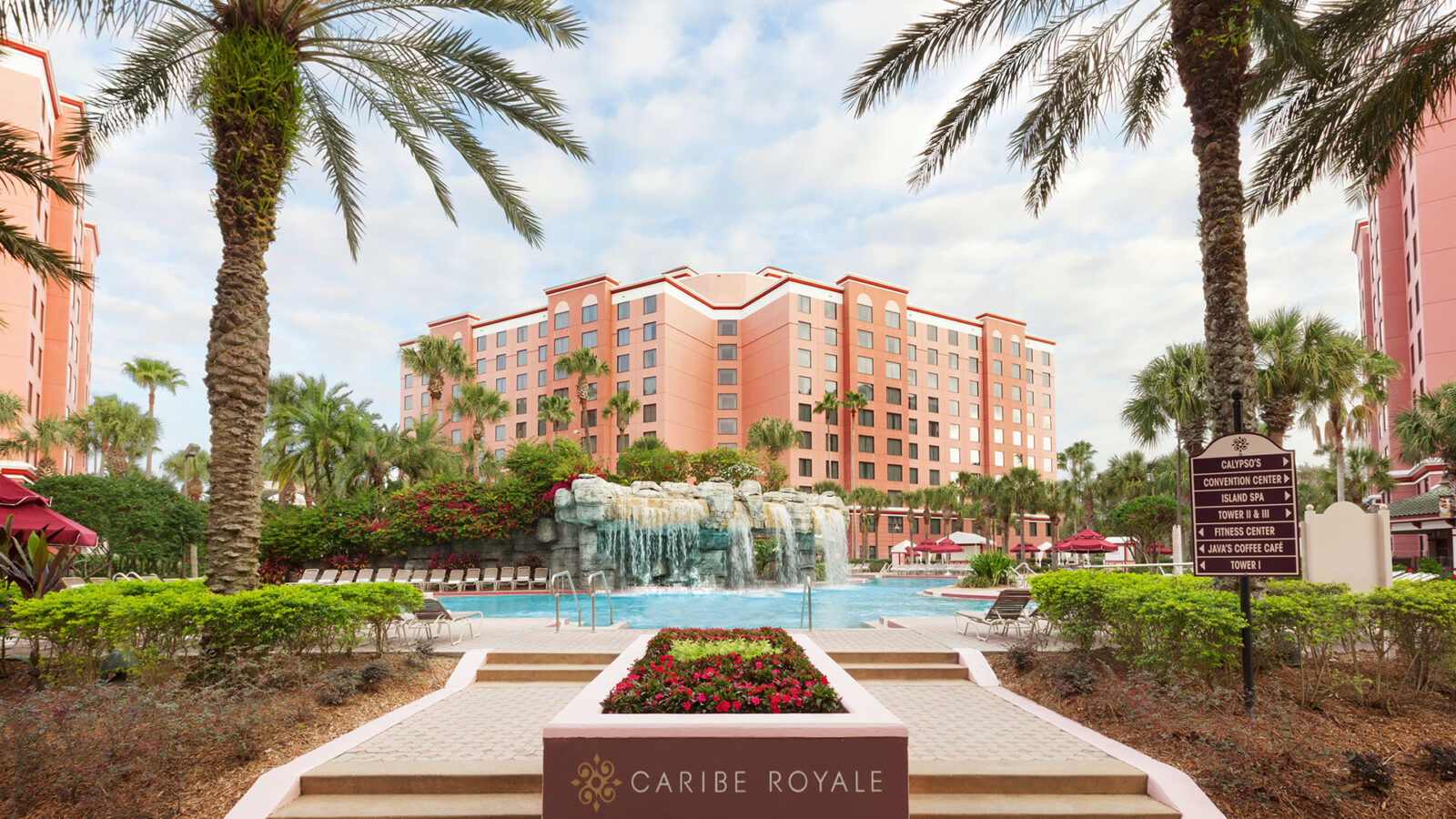 Pool with waterfall feature framed by two palm trees, and pink hotel towers in background