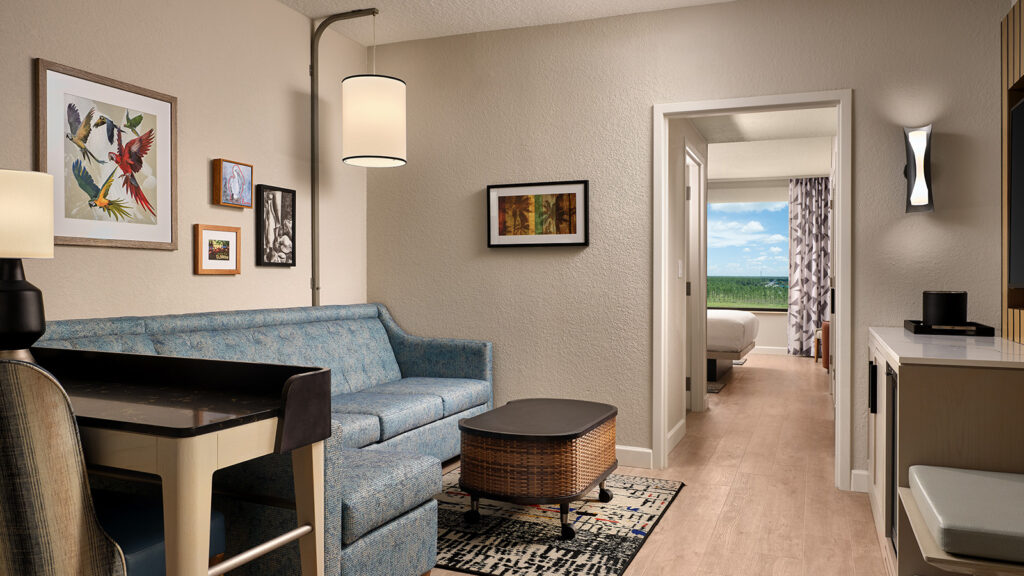 A hotel room with a sofa bed and table that leads into a bedroom with a large window