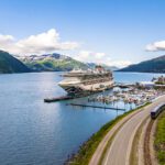 Cruise ship docked in bay alongside a bluff with a train going by and mountains in the background
