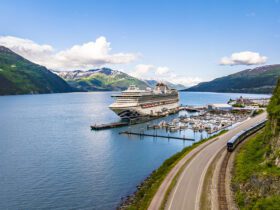 Cruise ship docked in bay alongside a bluff with a train going by and mountains in the background