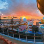 Cruise ship deck with water slides and a ropes course at sunset, with city skyline in background