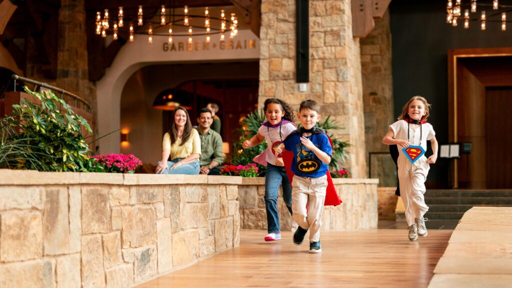Three kids in super hero caps running down a wood floor at a hotel