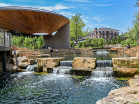 People hanging out at pond with tiered waterfall feature in park with large pavilion and buildings in background