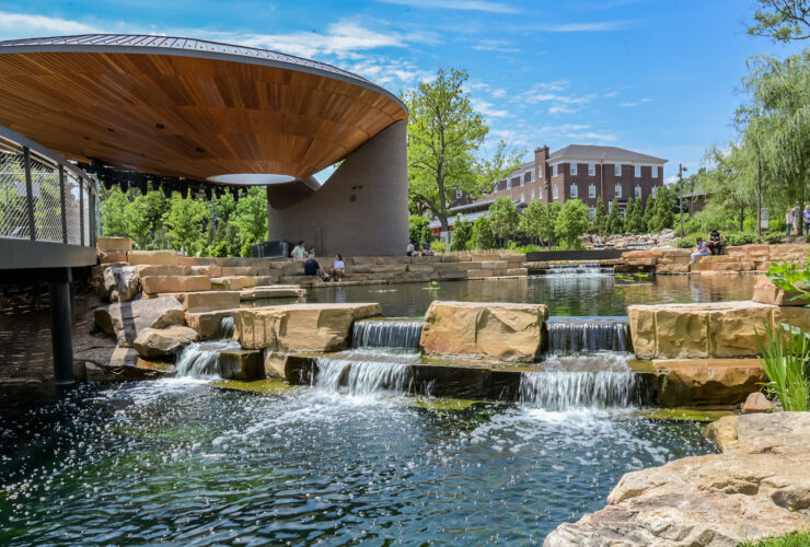 People hanging out at pond with tiered waterfall feature in park with large pavilion and buildings in background