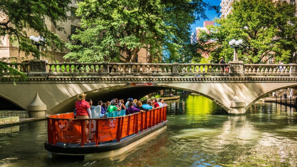 A boat filled with people about to go under a bridge on the San Antonio River Walk