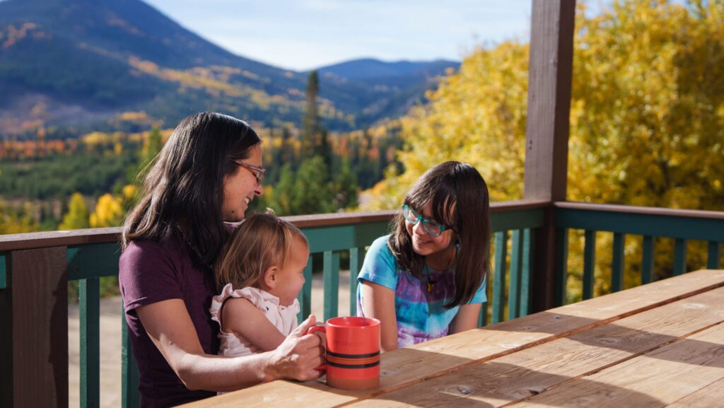 A woman with two kids sitting on the outdoor deck of a cabin at YMCA of the Rockies with the mountains behind them